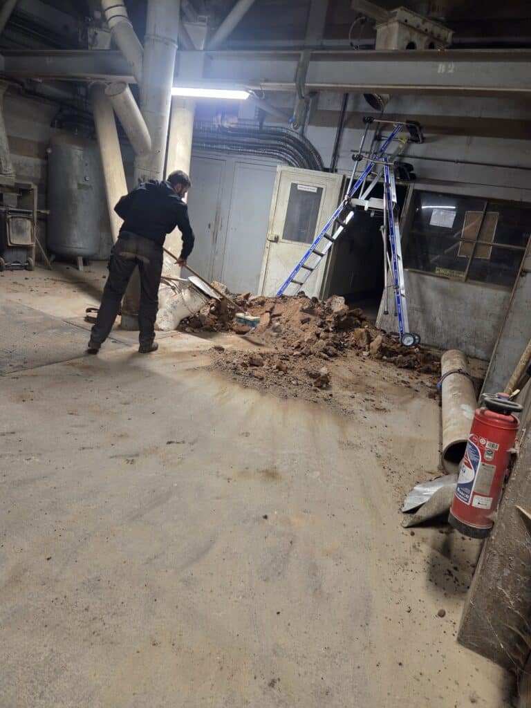 Ouvrier pelletant des gravats dans une usine. Panneau Défense de fumer visible en arrière-plan.