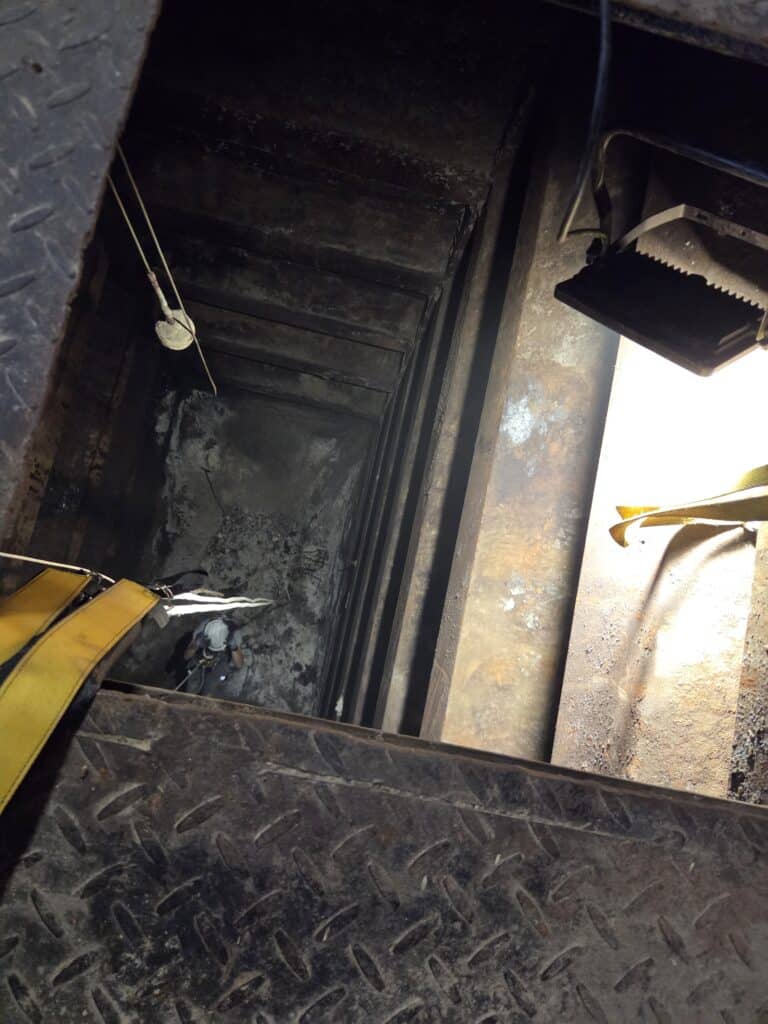 Ouvrier en harnais descendant dans un puits industriel sombre via une trappe métallique éclairée.