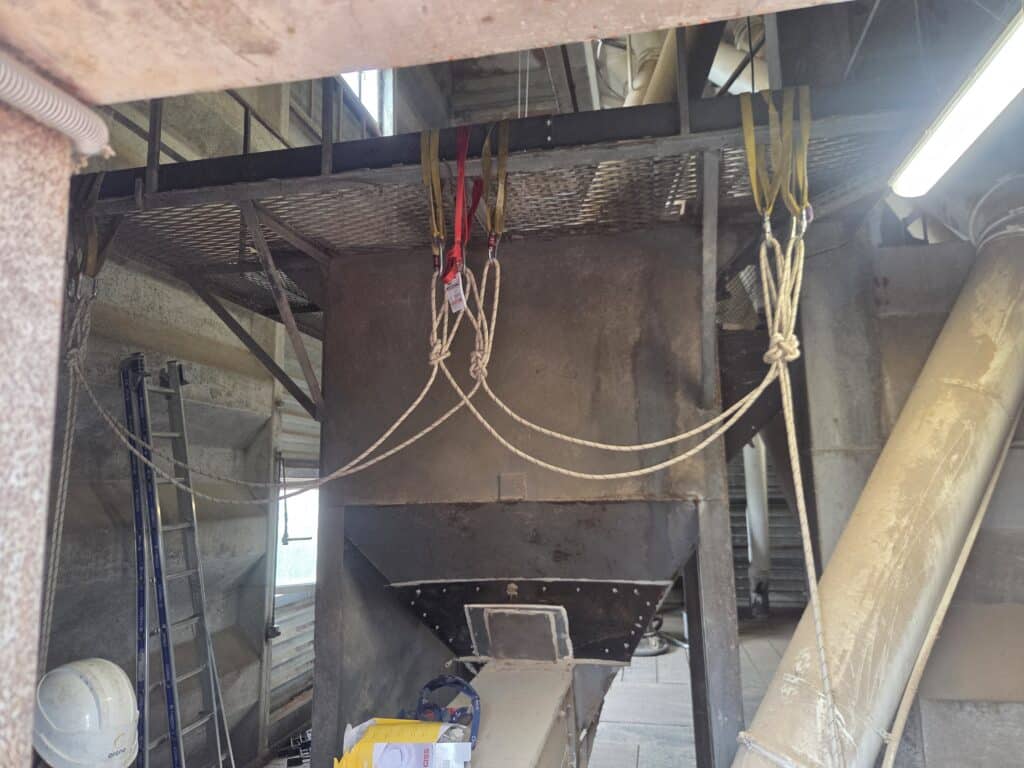 Trémie industrielle suspendue par cordes et sangles, avec échelle et casque Orano dans une usine.